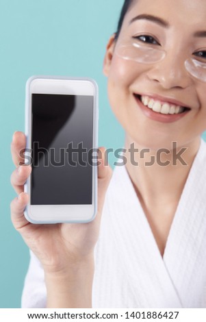 Cropped photo of charming happy female model in white bathrobe with fresh pure skin standing against blue background. She is holding smartphone in arms and showing screen to camera. Cosmetology
