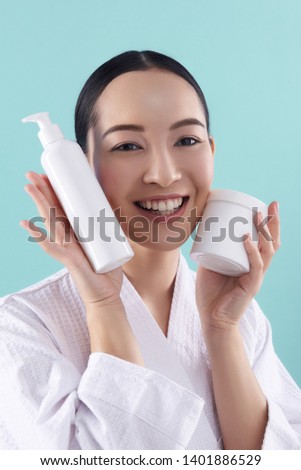 Close up of happy lady holding white bottles with cosmetics in arms against blue background in studio. Cosmetology, beauty and spa concept. Copy space on bottle