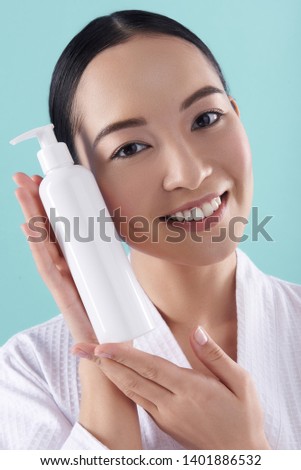 Close up of smiling lady holding white bottle with cosmetics and posing for camera against blue background. Cosmetology, beauty and spa concept. Copy space on bottle