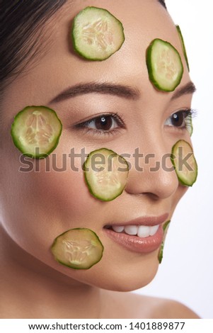 Calm young Asian lady smiling and looking into the distance while having cucumber slices on her face
