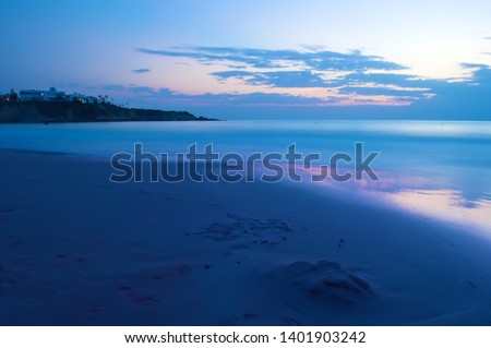 View of empty sandy Coral Bay beach near Paphos, Cyrpus. Sunset, orange sky above dark blue shallow water with waves. Warm evening in fall. Copy space. Long exposure