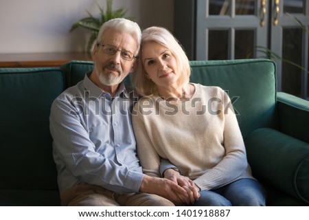Serious hoary couple of retirees hugging holds hands symbol of devotion and care, spouses sitting on sofa look at camera, forever love, medical insurance, nursing home, services for old people concept