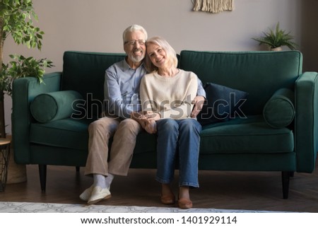 Grey haired couple in love embracing sitting on couch smiling looking at camera feels healthy, harmony in relationships, happy long life together, anniversary, older people services insurance concept