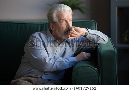 Sixty years old mature man sitting alone on couch at home thinking lost on sad thoughts, pondering about personal or health problems and midlife crisis, senior widower male feels lonely concept image Royalty-Free Stock Photo #1401929162
