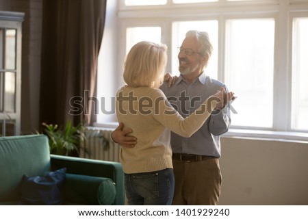Happy active sixty years old spouses standing in living room moving, grey-haired husband embraces blond wife aged couple dancing waltz celebrating life event, romantic mood dating of retirees concept Royalty-Free Stock Photo #1401929240