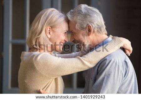 Aged cheerful couple standing indoors embracing looks at each other with love and devotion, close up side faces blond wife cuddles hoary husband old spouses touch foreheads tender moment concept image Royalty-Free Stock Photo #1401929243