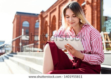 Beauty girl is sitting on a stairs and enjoy something on tablet computer. Red brick walls of building on the background.