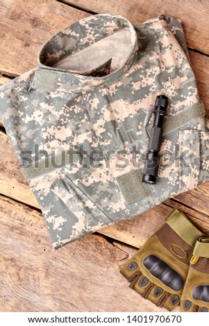 Set of military outfit on brown wooden background, flat lay. Folded camouflage, torch and fingerless gloves.