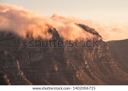 Landscape photo of rocky mountains with steep cliffs with their peaks partially covered with clouds at sunset. Pictured : The Twelve Apostles, shot from Lion's Head in Cape Town, South Africa.  