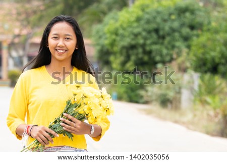 Young beautiful Asian women with happy big smile on countryside background. She got dark tan skin with long hair and holding flowers walking on the street with yellow cloth traditional thai style