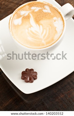 Delight in the artful presentation of a cappuccino intricately adorned accompanied by a tantalizing chocolate heart showcased on a lustrous polished tabletop
