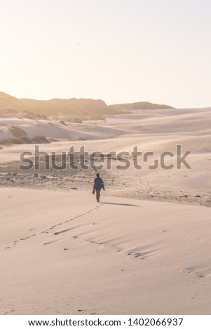 Portrait photo of a lonely woman walking barefoot on a huge, windy beach with sand dunes at sunset. Shot in Platboom beach, Cape Town, Western Cape, South Africa.