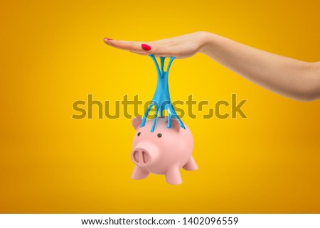 Pink piggy bank stuck to female hand with blue sticky slime on yellow background. Objects and materials. Banking and financial industry. Management and savings.