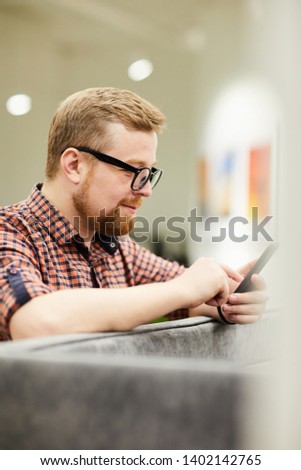Content pensive young bearded man in eyeglasses leaning on sofa and reading message on phone while communicating online in art museum
