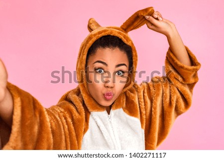 Young woman in bunny kigurumi standing isolated on pink background taking selfie picture on smartphone holding rabbit ear posing to camera pouting lips playful close-up.
