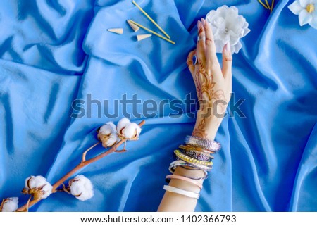 Slender female hand painted with Indian oriental mehndi ornaments by henna. Hand dressed in bracelets and rings hold white flower. Blue fabric with folds and cotton branches on background.