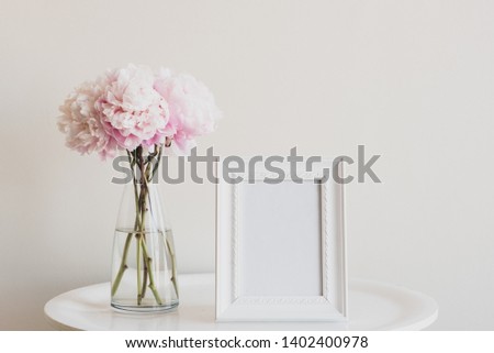 Close up of pale pink peonies in glass vase with blank rectangular picture frame on white table - matte filter effect and selective focus