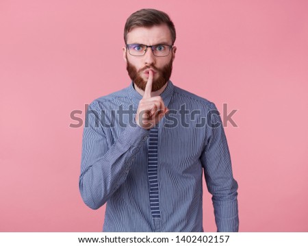 Young handsome red bearded man with glasses and a striped shirt, keeps finger on lips, tells secret information, demonstrates hush gesture isolated over pink background.