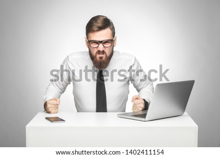 Portrait of aggressive angry young boss in white shirt and black tie are sitting in office and having bad mood,punch the table and clenching teeth. Indoor, studio shot, isolated, gray backgroud