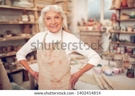 So happy. Joyful grey haired woman smiling to you while standing in her workshop