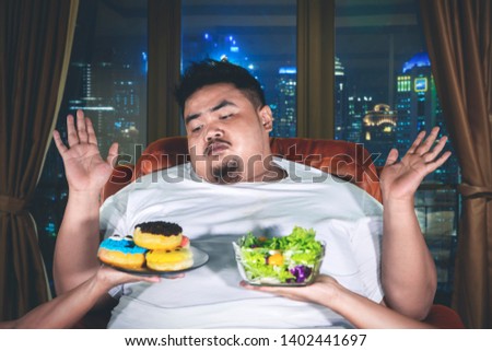 Picture of fat man choosing a vegetable salad or donuts with confused expression in the apartment