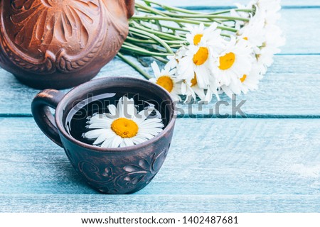 A teapot and a cup of tea stand on wooden blue boards with daisy flowers. Close-up.