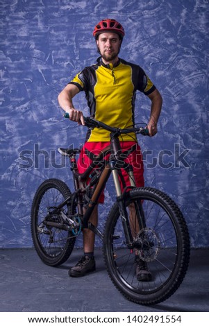 Cyclist in a yellow T-shirt and red shorts is holding a mountain bike against the background of a blue wall. Full suspension. Active lifestyle, cycling.