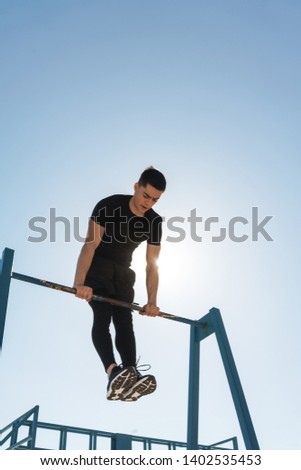 Photo of young sportsman 20s in black tracksuit doing acrobatics on horizontal gymnastic bar during morning workout by seaside