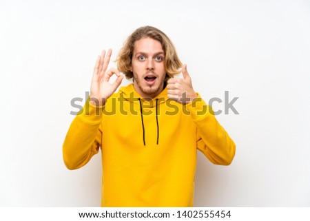 Blonde man with  sweatshirt over white wall showing ok sign and thumb up gesture
