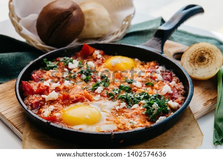 Mexican scrambled eggs with two eggs and sauce and tomatoes, red sweet pepper and chili pepper with beans, decorated with finely chopped dill on a large black pan on a wooden иoard