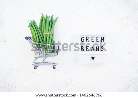 Creative scandinavian style flat lay top view of fresh green beans in shopping cart and text in lightbox on concrete table background copy space. Minimal house cooking concept promo template