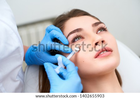 Doctor in medical gloves with syringe injects botulinum under eyes for rejuvenating wrinkle treatment. Filler injection for eye wrinkles smoothing. Plastic aesthetic facial surgery in beauty clinic Royalty-Free Stock Photo #1402655903