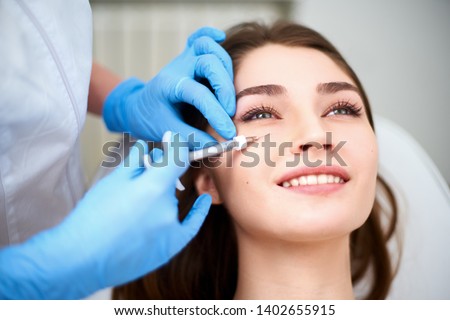 Doctor in medical gloves with syringe injects botulinum under eyes for rejuvenating wrinkle treatment. Filler injection for eye wrinkles smoothing. Plastic aesthetic facial surgery in beauty clinic Royalty-Free Stock Photo #1402655915