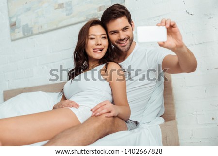 selective focus of happy man taking selfie with girlfriend showing tongue in bedroom 