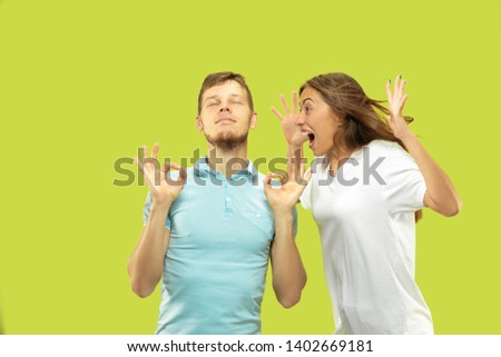 Beautiful young couple's half-length portrait isolated on green studio background. Man is trying to keep calm with closed eyes while woman is screaming. Facial expression, human emotions concept.