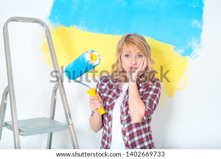 Tired woman ready to makes repairs at home. Sad woman with paint roller, brush and ladder. Girl painting big paint Ukrainian flag on wall at home. Concept of home repair and design.