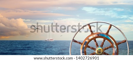 Marine landscape with a steering wheel and sailing yacht on skyline. Calm sea with a cloudy sky at sunrise and flying seagulls for your concept about romantic sea voyage on a ship around the world. Royalty-Free Stock Photo #1402726901