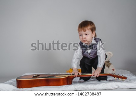 cute Caucasian boy in bow tie and vest with acoustic guitar on grey background