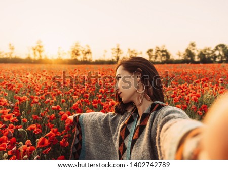 Point of view image of beautiful young woman making selfie in poppy flower field at sunset in summer outdoor.