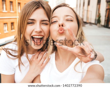 Two young smiling hipster blond women in summer clothes. Girls taking selfie self portrait photos on smartphone.Models posing on street.Female making duck face and shows peace sign