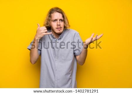 Blonde man over isolated yellow background making phone gesture and doubting