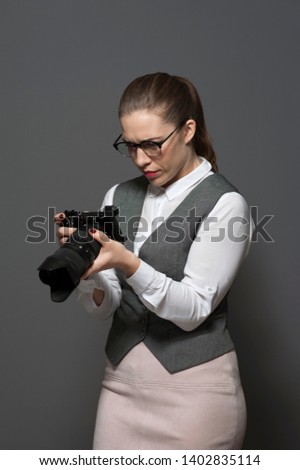 Portrait of young female photographer in eyeglasses dressed in casual clothes holding dslr photo camera standing on grey background