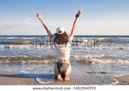 Young woman in swimsuit and white t-shirt with her hands raised is feeling happy and freedom, relaxing, sitting on the sand, looking at sea. Vacation and travel concept.