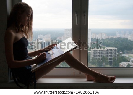 Silhouette of young woman is drawing a watercolor painting. The female artist paints watercolour landscape. Teen girl with blond hair is sitting on the window sill with brush in hand. Creative concept