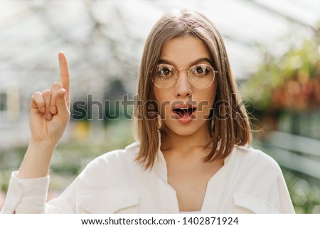 Woman in shirt and glasses has idea. Photo of amazed girl posing with finger up on blur background.