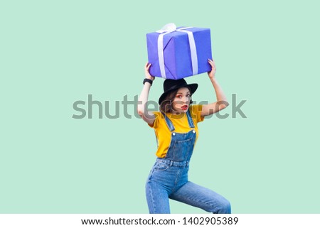 Suprised funny young girl in hipster wear in denim overalls and black hat standing and holding under head giant big heavy gift box with unbelievable face. Studio shot, green background, isolated