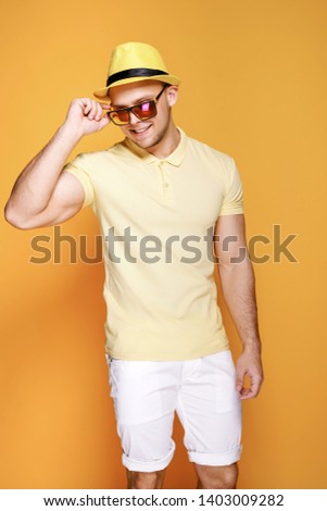 Positive young male in trendy outfit - yellow shirt, sunglasses, white shorts, holding straw hat smiling and looking away while standing against yellow background