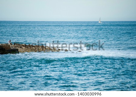 beach waves and breakwater rocks break strong with white foam and man takes pictures with a sailboat in the backgroun