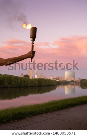 Flaming torch held high over Indianapolis skyline symbolizing enlightenment, hope, life, truth, and regenerative power of flame.  The torch reflects the light of knowledge, which dispels the darkness