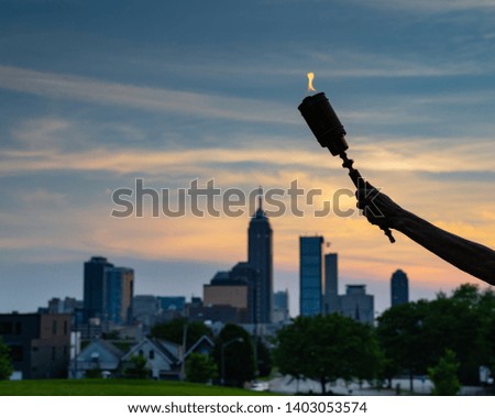 Flaming torch held high over Indianapolis skyline symbolizing enlightenment, hope, life, truth, and regenerative power of flame.  The torch reflects the light of knowledge, which dispels the darkness